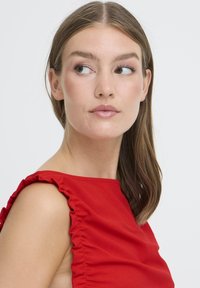 Top rojo sin mangas con detalles de volantes en los hombros, textura suave y diseño ajustado, presentado sobre un fondo blanco liso.