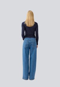Jeans in denim blu a gamba larga con vita alta, dotati di tasche posteriori e chiusura con zip. Il modello indossa un top aderente a maniche lunghe.