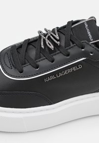 Musta nahkainen lenkkari, jossa on teksturoitu pinta, valkoinen kumipohja, koristeelliset mustat nauhat ja hopeanvärinen logoyksityiskohta sivulla.