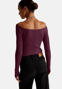 Schulterfreies geripptes Oberteil in tiefem Lila, mit langen Ärmeln und figurnaher Passform. Kombiniert mit hochtaillierten schwarzen Jeans und einem Leder-Tag.