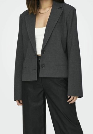Femme portant un blazer court gris foncé sur un crop top blanc avec un pantalon noir taille haute à jambes larges et un collier chaîne doré.