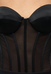 Μαύρο κορσές χωρίς τιράντες με διαφανείς μεσενδεριές και κάθετες λεπτομέρειες κορσέ, με ένα μικρό χρυσό κούμπωμα στο κέντρο μπροστά.