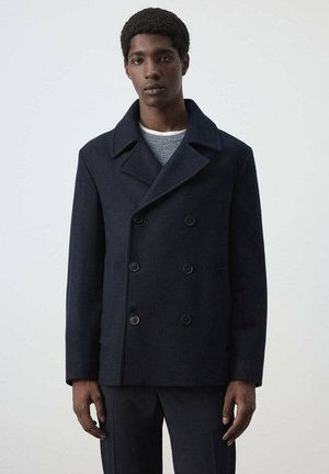 Un homme porte un manteau croisé bleu marine foncé sur un pull gris et pose devant un fond uni clair.