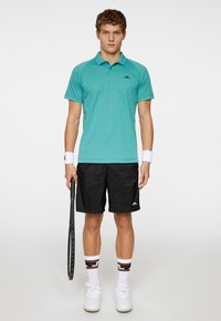 Turkos polo skjorta med en svart logotyp, tillsammans med svarta shorts. Vita sportstrumpor med färgglada detaljer och vita sneakers. Håller en tennisracket.