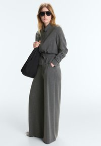 Femme portant un costume gris à fines rayures avec ceinture et pantalon à jambes larges, tenant un sac noir sur l'épaule et portant de grandes lunettes de soleil noires.