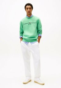 Sweat-shirt vert avec le logo "TOMMY HILFIGER EST. 1985", porté par-dessus une chemise rayée bleue et blanche, associé à un pantalon blanc et des baskets claires.