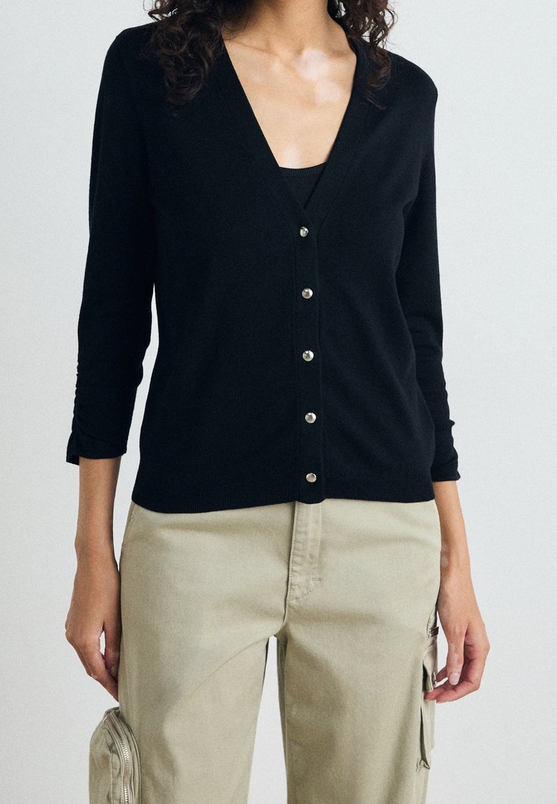 Svart kortärmad cardigan med V-ringning, sju knappar framtill och ärmar i trekvarts längd, kombinerad med ljusbeiga cargobyxor.