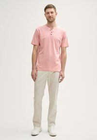 Mand står op, iført en lys pink kortærmet henley skjorte, beige bukser og hvide sneakers foran en ensfarvet hvid baggrund.