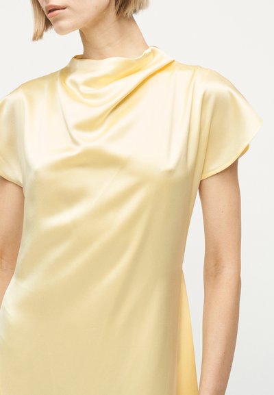 Γυναίκα που φοράει φόρεμα από απαλό κίτρινο σατέν με ντραπαρισμένο ντεκολτέ και κοντά μανίκια, εμφανιζόμενη από τους ώμους έως τη μέση.
