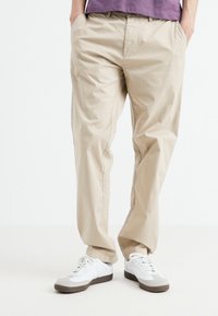 Beige cotton blend trousers with a relaxed fit, featuring a waistband and side pockets, paired with white and gray sneakers.