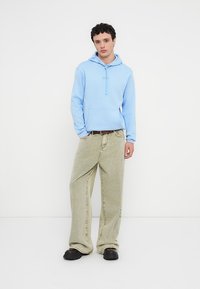 Ανοιχτό μπλε βαμβακερό φούτερ με κουκούλα, τσέπη καγκουρό και κορδόνι, συνδυασμένο με φαρδιά παντελόνια κοτλέ σε ελιά πράσινο χρώμα και μαύρα παπούτσια.