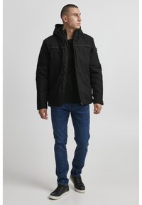 Svart isolert jakke med hette, med glidelås og to sidelommer, sammen med blå jeans og svarte sneakers.