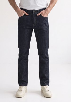 Mand iført mørkeblå jeans med lige ben, hænderne i lommerne og hvide sneakers, stående mod en ensfarvet lys baggrund.
