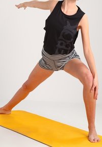 Black sleeveless top with a graphic print, striped shorts, and a yellow yoga mat. The subject is in a warrior pose. Smooth background.