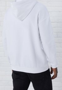 Vista trasera de una persona que lleva una sudadera blanca lisa y jeans negros, con un fondo de pared de ladrillo blanco.