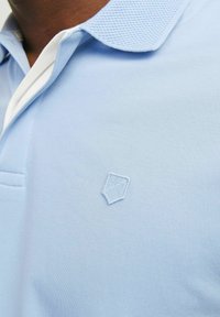 Világoskék póló ing fehér gallérral, texturált gallér anyaggal és egy kis hímzett logóval a mellrészen. Finom kötött textúrájú az egész.