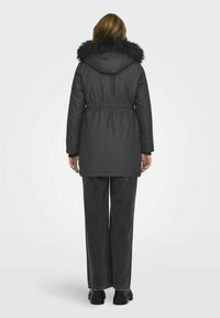 Parka rembourrée gris foncé avec une capuche en fourrure, taille élastique, manches longues et poches latérales, associée à un jean noir à jambes larges et des bottes imposantes.