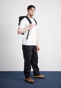 Mochila negra, camiseta blanca con texto, pantalones negros y botas de senderismo marrones. Tejido suave en la mochila, diseño robusto, colores neutros.