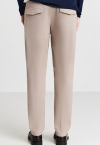 Pantaloni beige con una texture liscia, design affusolato e due tasche posteriori. Indossati con scarpe scure su uno sfondo chiaro.