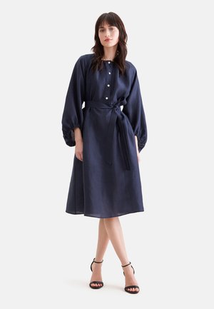Femme portant une robe bleu marine au genou avec des manches longues bouffantes, des boutons sur le devant, une ceinture nouée à la taille et des talons noirs à bride à la cheville, debout.