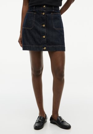 Mørk denim nederdel med knapper foran og forlommer, båret med sorte loafers på en person, der står mod en hvid baggrund.