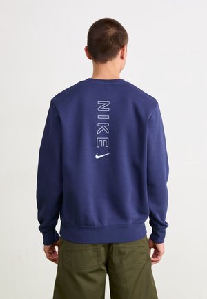 Mand iført marineblå Nike-sweatshirt med vertikalt hvidt logo på ryggen, kombineret med olivengrønne bukser, står mod en ensfarvet baggrund.
