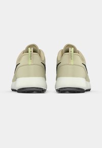 Τα μπεζ παπούτσια γκολφ διαθέτουν λείο επάνω μέρος, μαύρες λεπτομέρειες και μαλακή λευκή σόλα με υφασμένο μαύρο πέλμα για πρόσφυση.