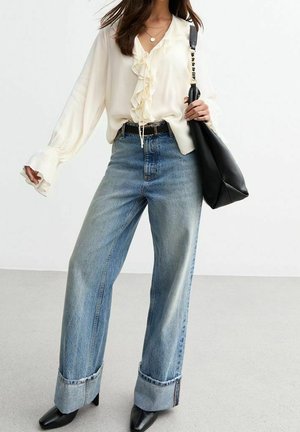 Vrouw draagt een crèmekleurige blouse met ruches, wijde jeans met manchetten, zwarte puntige schoenen en draagt een grote zwarte schoudertas.