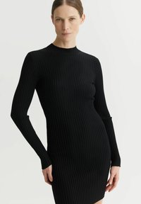 Czarna dzianinowa sukienka z prążkowanej tkaniny, z długimi rękawami i wysokim dekoltem; dopasowany krój z bocznymi wycięciami w okolicy mankietów i lekko rozkloszowanym dołem.