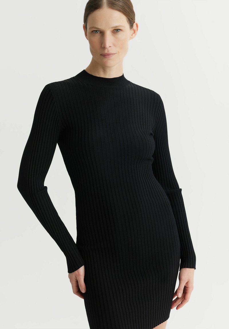 Czarna dzianinowa sukienka z prążkowanej tkaniny, z długimi rękawami i wysokim dekoltem; dopasowany krój z bocznymi wycięciami w okolicy mankietów i lekko rozkloszowanym dołem.
