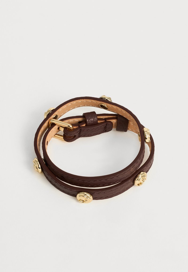 Bracelet enveloppant en cuir marron avec des clous ronds dorés et une boucle dorée, enroulé soigneusement sur un fond clair et uni.