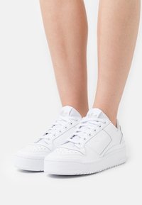Vita lädersneakers med perforerat tåsidas, tjock vit sula och strukturerade detaljer. Innehåller en logotyp på sidan och snörningsdesign.