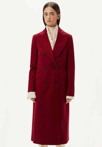 Dubbelknäppt frack i djupröd ull, med svarta knappar, stora slag och långa ärmar med vita strukturerade manschetter. Lång silhuett.