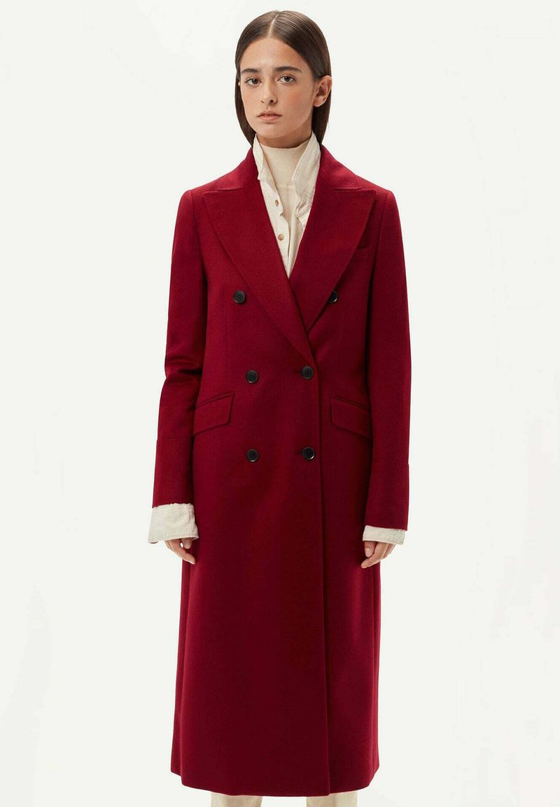 Dubbelknäppt frack i djupröd ull, med svarta knappar, stora slag och långa ärmar med vita strukturerade manschetter. Lång silhuett.