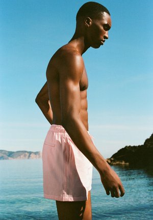 Ing nélkül áll egy rózsaszín fürdőnadrágot viselő férfi a nyugodt tengerpart mellett tiszta ég alatt, sziklás partvonal a háttérben, lefelé néz.