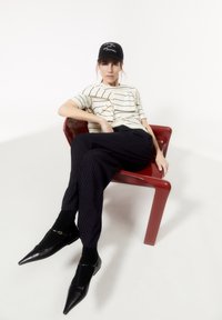 Model zittend op een rode plastic stoel, gekleed in een gestreepte beige top, donkergestreepte broek en zwarte puntige schoenen met een gesp.