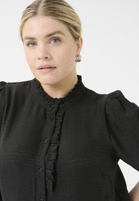 Černá, strukturovaná blůza s nabíranými rukávy a volánkovou légou. Uprostřed má knoflíkové zapínání. Viditelné stříbrné náušnice.