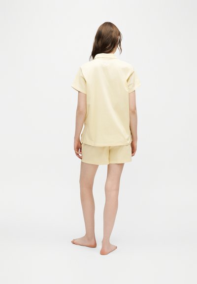 Femme debout pieds nus, de dos, portant un t-shirt jaune pâle à manches courtes et un short assorti sur un fond blanc uni.