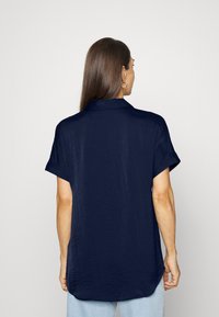 Camicetta corta a maniche corte di colore blu scuro, con colletto, vestibilità rilassata, tessuto testurizzato e orlo arrotondato. Vista posteriore che mostra una leggera drappeggio.