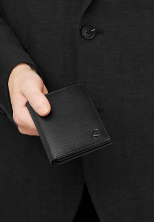 Ruka držící černou texturovanou koženou peněženku na pozadí tmavého sakového saka s knoflíky.