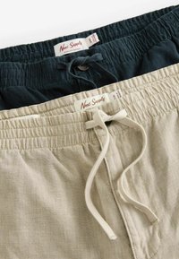 Two pairs of lightweight shorts featuring an elastic waistband, drawstring ties, one in dark green and the other in beige. Labels visible.