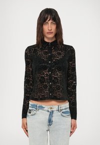 Woman wearing a black floral lace button-up top with collar and light-wash high-waisted jeans against a white background.