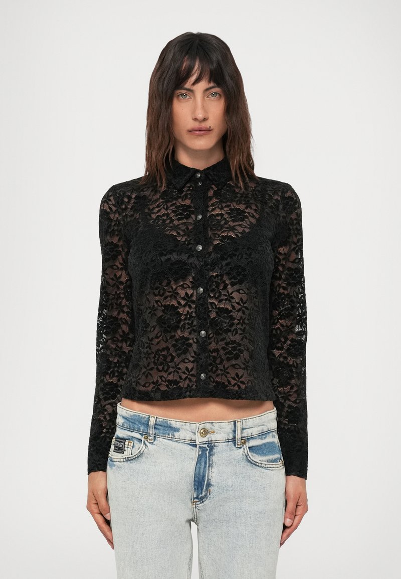 Mulher a usar uma blusa preta de renda floral com botões e colarinho, combinada com jeans de cintura alta em lavagem clara, contra um fundo branco.