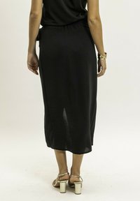 Jupe midi noire avec une coupe décontractée, dotée d'une taille élastique froncée et d'une fente sur le côté, portée avec des sandales à talons blocs métalliques.