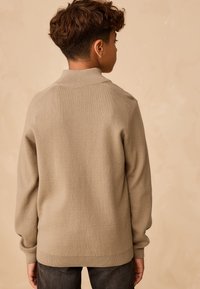Ragazzo con capelli ricci che indossa un maglione beige e pantaloni scuri, in piedi di spalle su uno sfondo neutro e semplice.