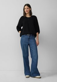 Czarny top o fakturowanej powierzchni z okrągłym dekoltem, luźny krój i rękawy o długości trzech kwart. W połączeniu z niebieskimi dżinsami typu flare o wysokim stanie i jasnym obuwiem.