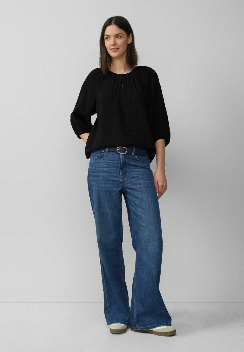 Czarny top o fakturowanej powierzchni z okrągłym dekoltem, luźny krój i rękawy o długości trzech kwart. W połączeniu z niebieskimi dżinsami typu flare o wysokim stanie i jasnym obuwiem.