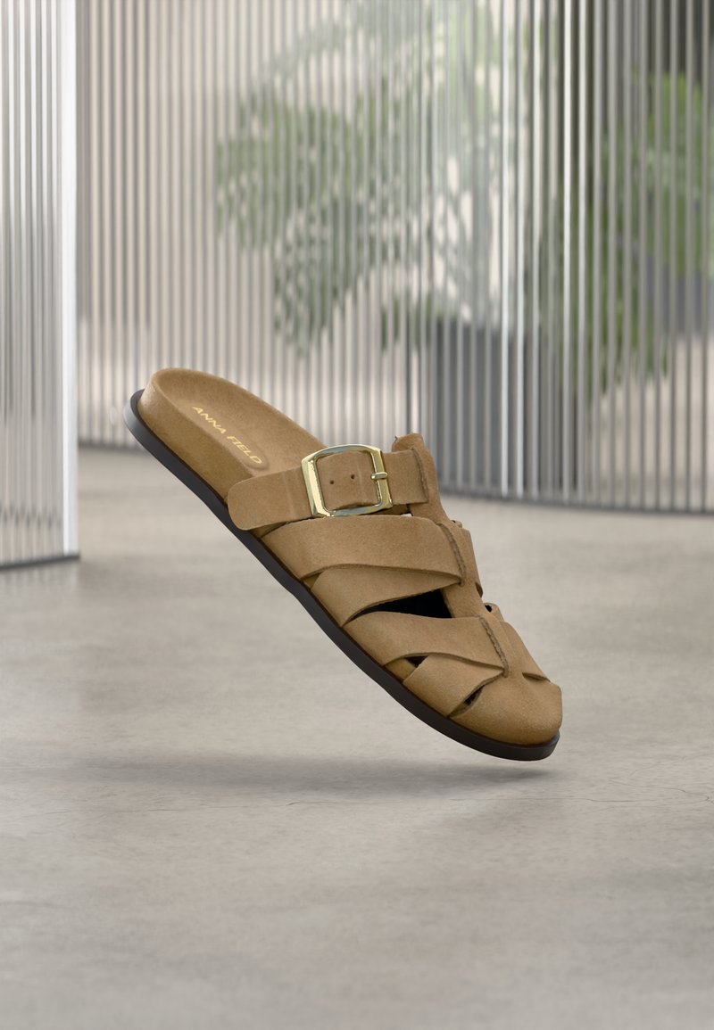 Sandale slide pour homme en daim beige avec boucle dorée, brides croisées et semelle noire, suspendue au-dessus d'un sol gris avec un arrière-plan flou.