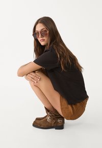 Czarny t-shirt, brązowe zamszowe szorty oraz brązowe zamszowe buty za kolano z klamrami. Modelka kuca, ma długie, faliste włosy i okulary przeciwsłoneczne.