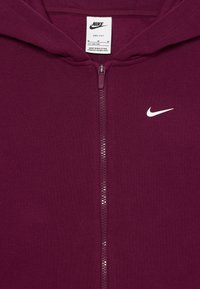 Sweatshirt de fecho marrom, feito de material Dri-FIT, com um logótipo branco da Nike no lado esquerdo do peito e um detalhe de fecho de malha.